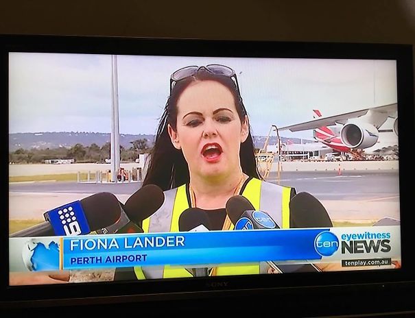 Woman at Perth Airport giving an interview, wearing a safety vest, with microphones around her. Airplane in background.