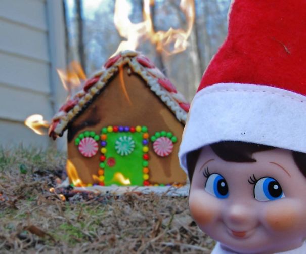 "Elf On The Shelf idea with the elf smiling mischievously in front of a gingerbread house that appears to be on fire in the background."