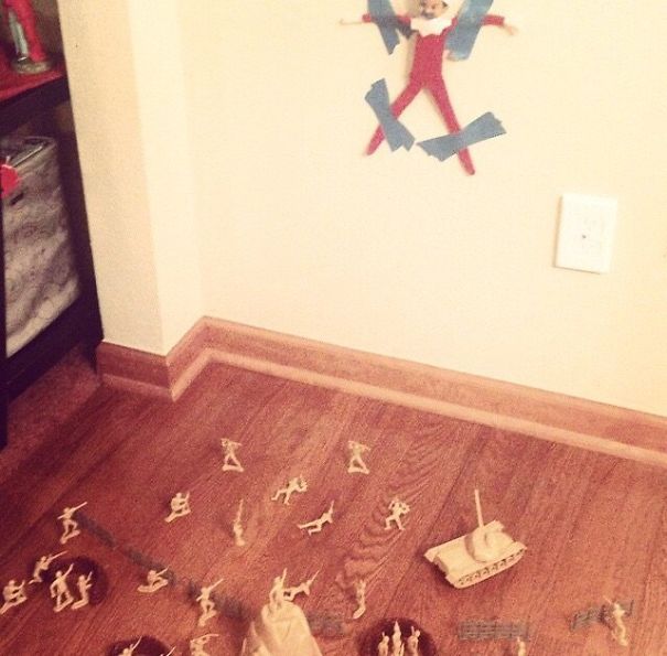 "Elf On The Shelf idea with the elf taped to the wall, surrounded by toy soldiers and a tank in a playful 'captured' military scene."