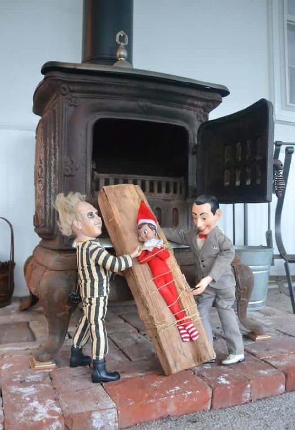  "Elf On The Shelf idea with the elf tied to a wooden plank, with Beetlejuice and Pee-wee Herman dolls appearing to place it into a wood stove."