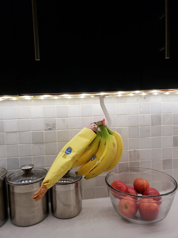 "Elf On The Shelf idea with elf hiding in a bunch of bananas, wrapped in a banana peel sleeve, creating a playful disguise in the kitchen."