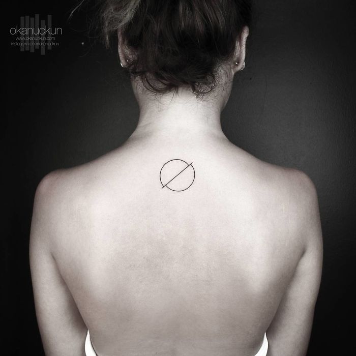 Geometric tattoo by Okan Uçkun, featuring a circle with a diagonal line, placed on the upper back against a dark background.
