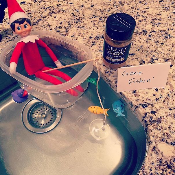  "Elf On The Shelf idea with an elf in a plastic container 'boat' fishing in the kitchen sink, surrounded by toy fish and a 'Gone Fishin’' sign."