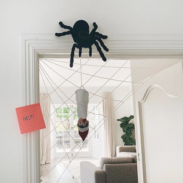 "Elf On The Shelf idea with the elf wrapped in webbing and hanging upside down in a doorway, caught in a spider's web with a 'HELP!' sign nearby."