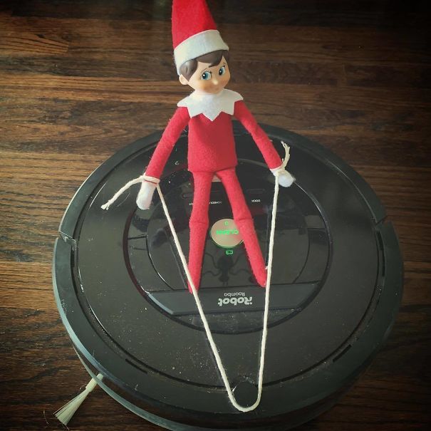 "Elf On The Shelf idea with elf posed as if skiing on top of a Roomba vacuum, holding strings like ski poles for a playful effect."