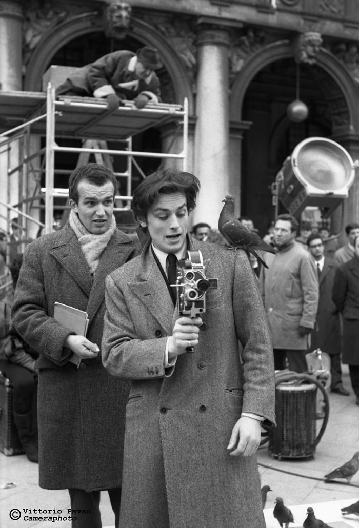 Two celebrities in Venice in the 50s filming with a vintage camera, surrounded by pigeons and onlookers.