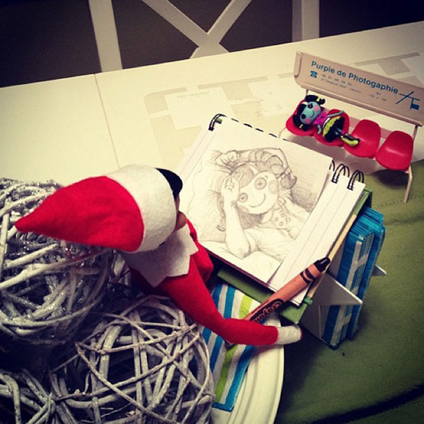 "Elf On The Shelf idea with the elf drawing a spooky sketch of a doll in a notebook, with a crayon in hand, creating an artist scene."