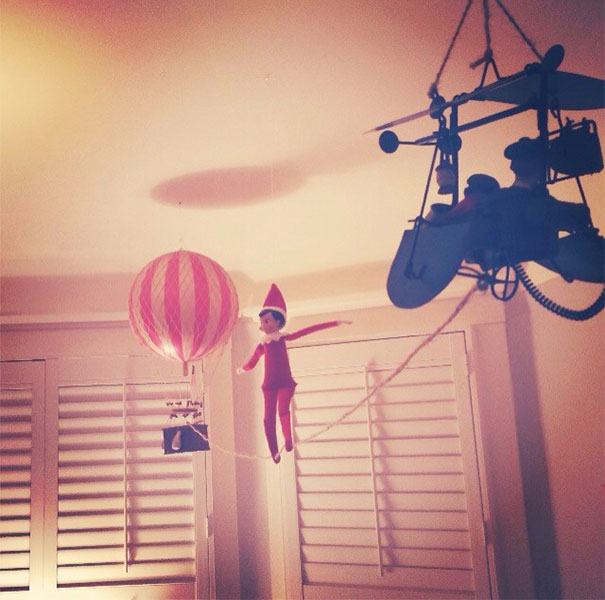 "Elf On The Shelf idea with the elf balancing on a tightrope between a hot air balloon and an airplane model, creating an adventurous scene."