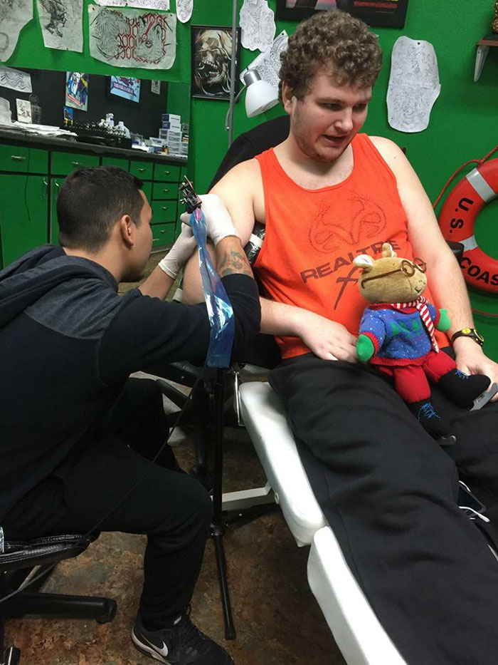 Autistic man receiving his dream tattoo in a shop after previous refusals, holding a teddy bear, wearing an orange tank top. Autistic man receiving his dream tattoo in a shop after previous refusals, holding a teddy bear, wearing an orange tank top.