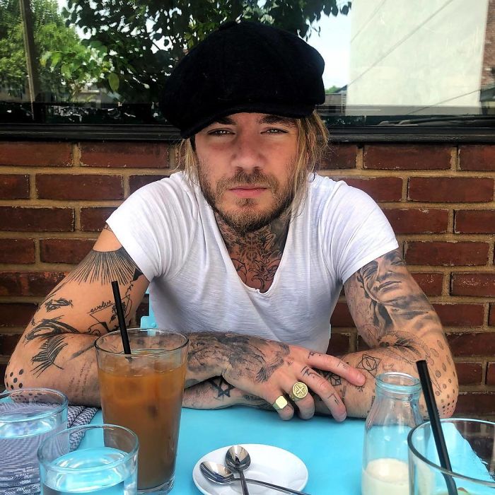 Tattoo artist with inked arms sitting at a table outdoors, wearing a black cap and white T-shirt. Tattoo artist with inked arms sitting at a table outdoors, wearing a black cap and white T-shirt.