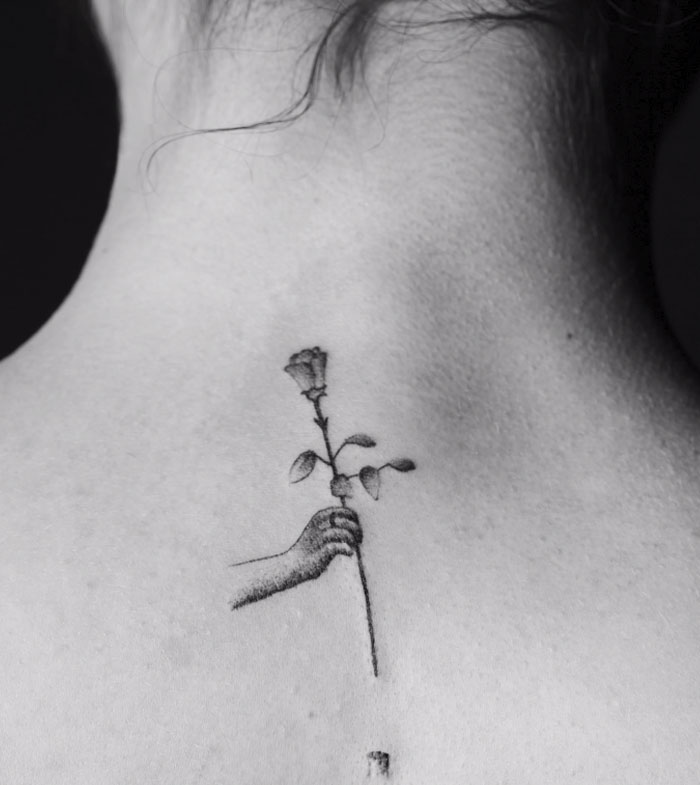 Tattoo of a hand holding a flower on a person's upper back, part of a motion tattoo series by an artist. Tattoo of a hand holding a flower on a person's upper back, part of a motion tattoo series by an artist.
