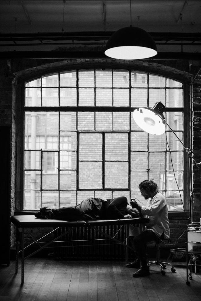 Artist inking a tattoo on a person in a studio with large industrial windows and overhead lighting. Artist inking a tattoo on a person in a studio with large industrial windows and overhead lighting.