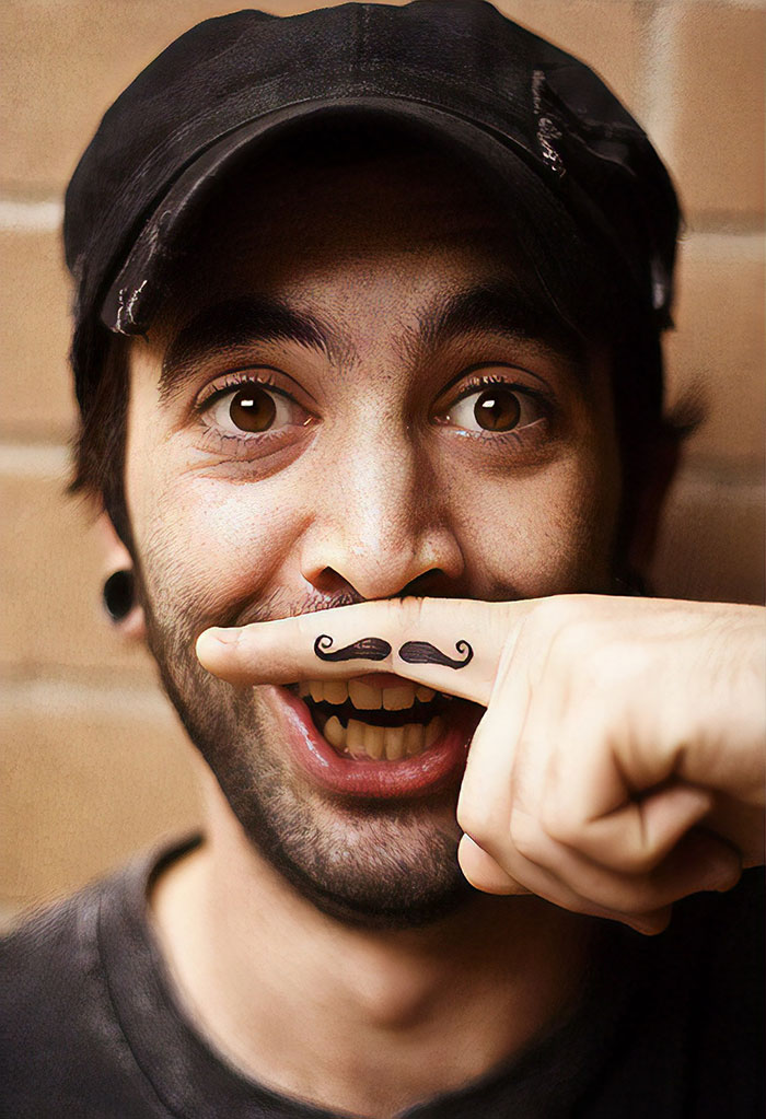 A person with a finger tattoo of a mustache, playfully holding it under their nose, smiling widely.
