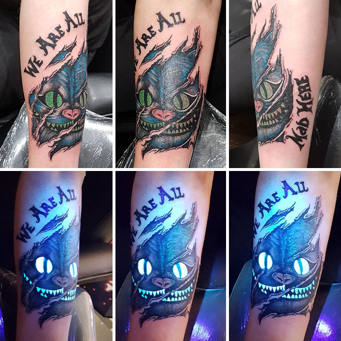 UV tattoo of a Cheshire Cat with glowing eyes and text "We Are All Mad Here" on an arm, vibrant under black light.