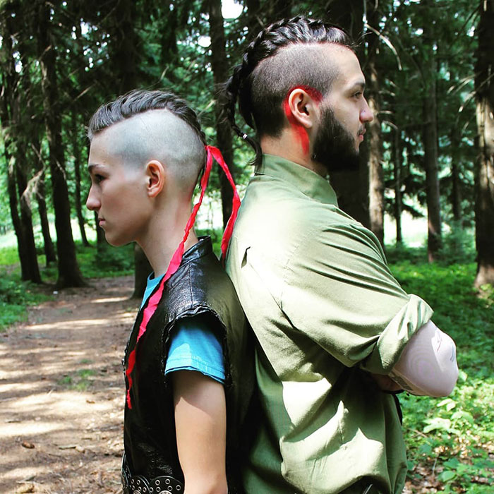 Two people with fierce Viking hairstyles stand back-to-back in a forest setting.
