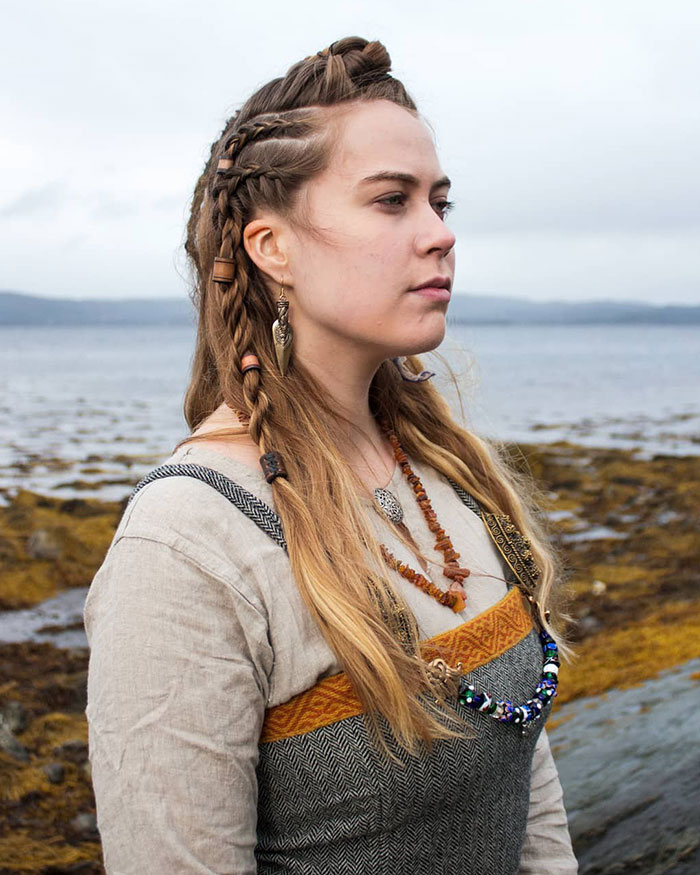 Woman with fierce Viking hairstyle, intricate braids and wooden beads, standing by the sea.