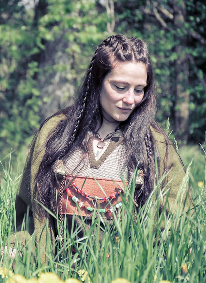 A woman with Viking hairstyle stands in a grassy field, wearing a traditional outfit, surrounded by nature.