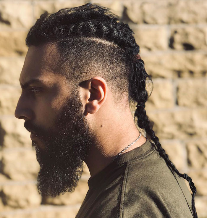 Man with a fierce Viking hairstyle, featuring braids and a neatly trimmed beard, against a stone wall background.