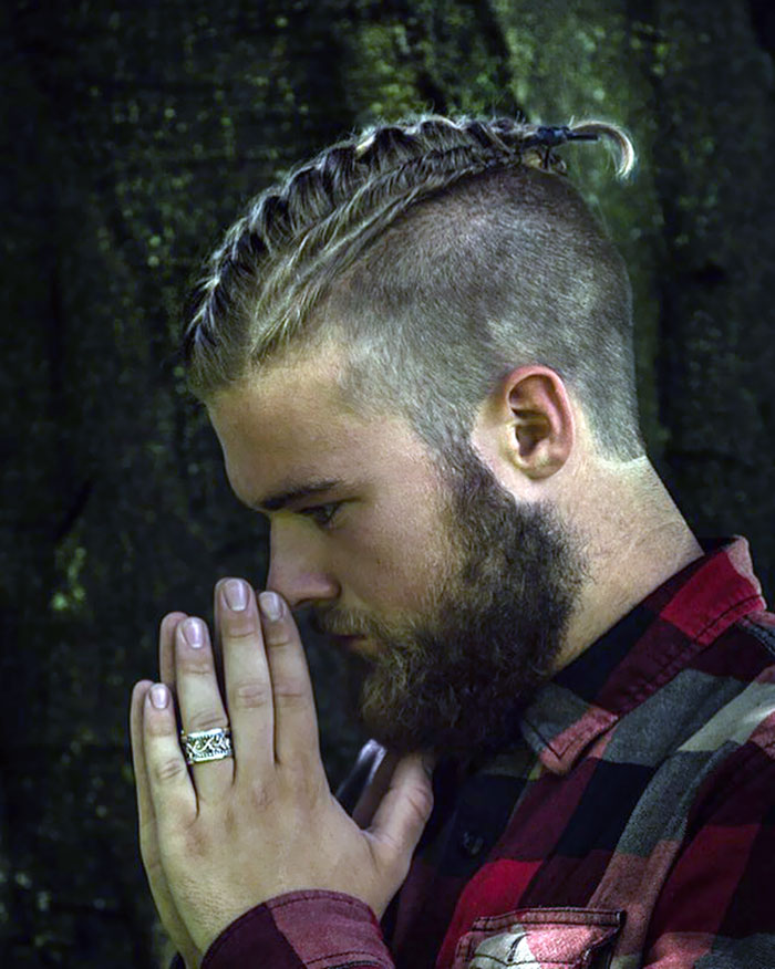 Man with braided Viking hairstyle, plaid shirt, and a ring, in deep contemplation outdoors.