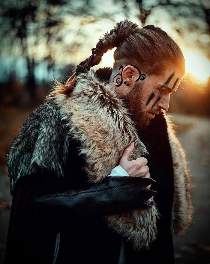 A man with fierce Viking hairstyle and braid, wearing fur, against a sunset backdrop.