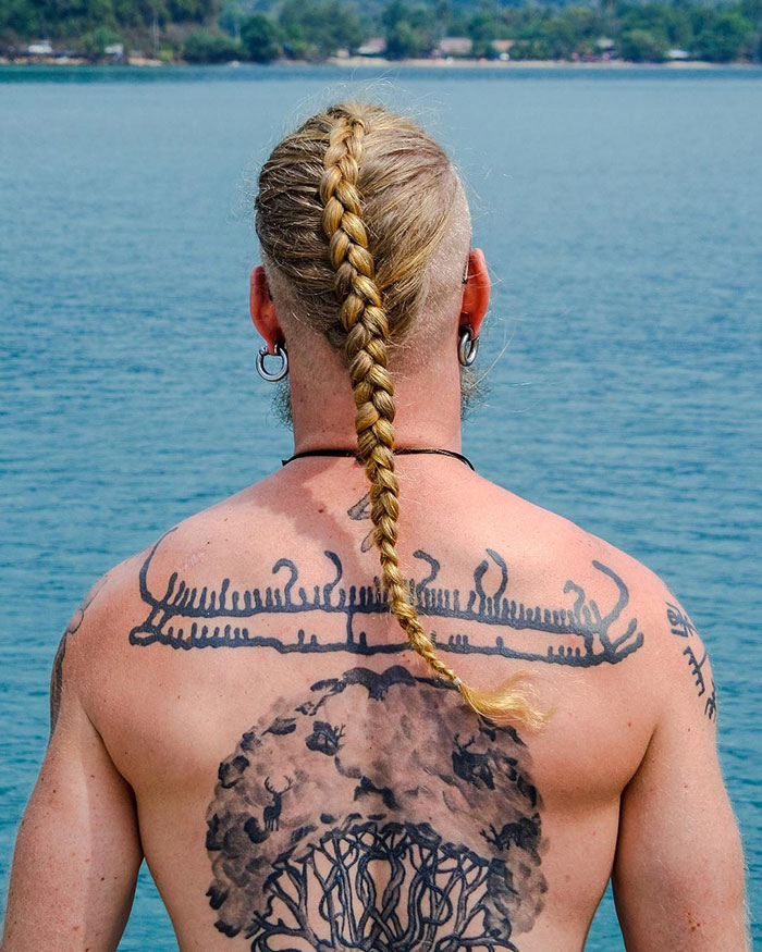 Blonde man with a braided Viking hairstyle and Norse tattoos overlooks a calm sea.