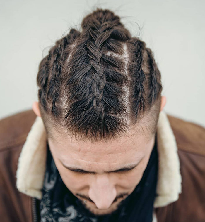 Man with intricate Viking hairstyle featuring tight braids and a bun, wearing a leather jacket with a shearling collar.
