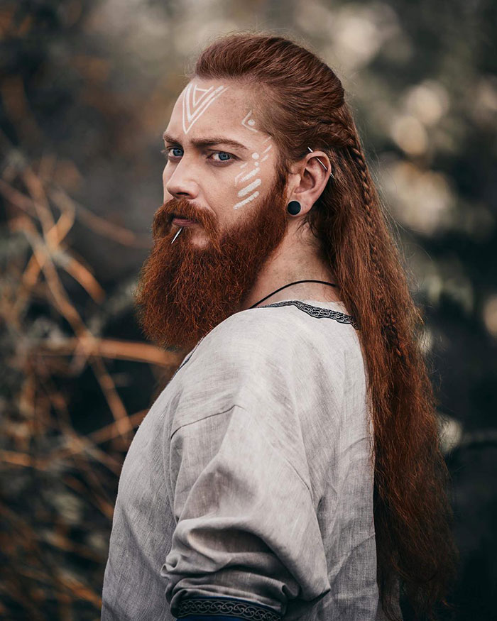 Red-haired man with fierce Viking hairstyle, long beard, tribal face paint, in a rugged outdoor setting.