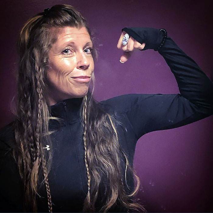 Woman showcasing Viking hairstyle, with long braids and fierce expression, flexing her arm against a purple background.