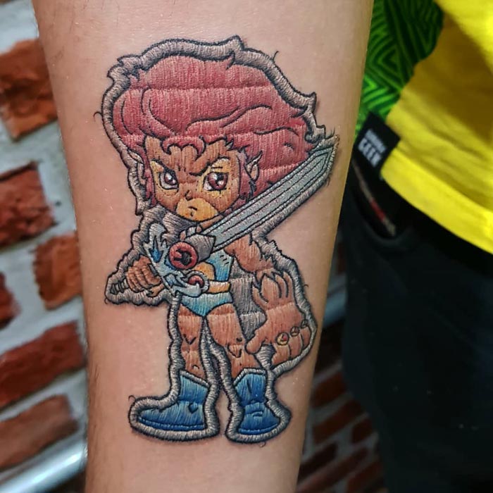 Embroidery tattoo on forearm featuring a cartoon character with a sword, created by a Brazilian tattoo artist.