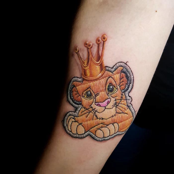 Embroidery tattoo of a lion cub with a crown on an arm, showcasing Brazilian tattoo artist's signature style.