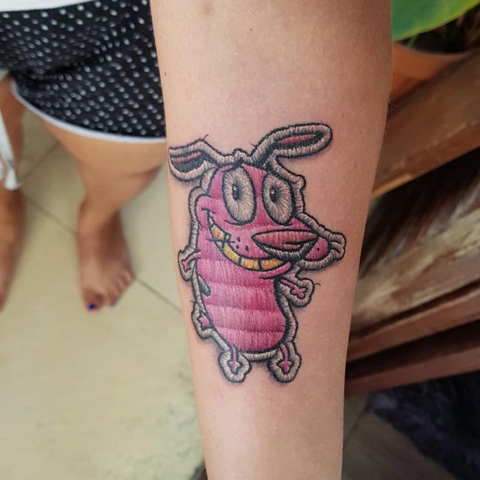 Embroidery tattoo of a pink cartoon dog on a forearm, showcasing detailed thread-like design by Brazilian artist.