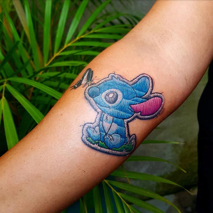 Embroidery tattoo of a blue cartoon mouse on an arm, showcasing intricate stitch-like design by Brazilian artist.