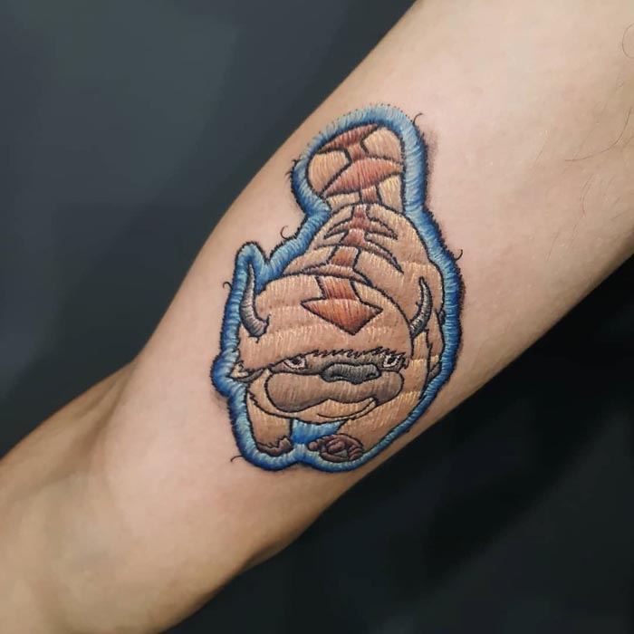 Embroidery tattoo of a cartoon creature on an arm, showcasing the Brazilian artist's unique style.