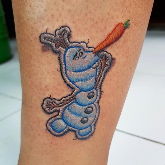 Embroidery tattoo of a cartoon snowman with a carrot nose on skin.