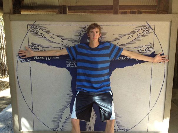 Tall young man in striped shirt showing arm span against a wall with a large Vitruvian Man drawing illustration.