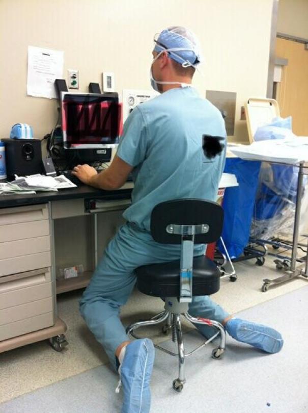 Tall person struggling to fit under a desk, sitting awkwardly on a small office chair in a medical setting.