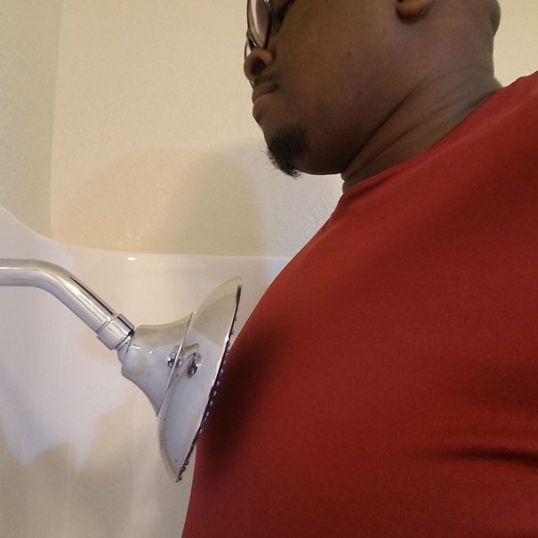 A tall person wearing glasses and a red shirt awkwardly fitting under a low showerhead in a bathroom.