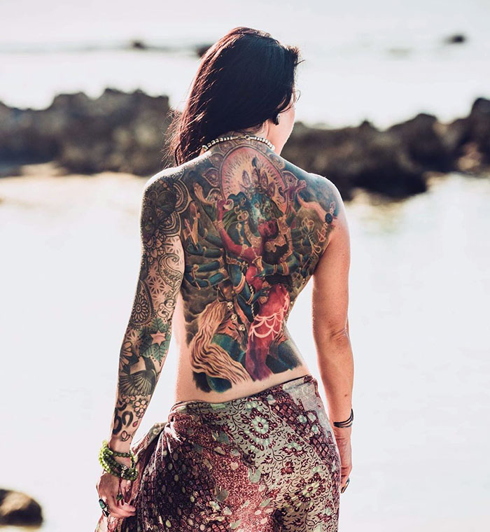 Person with an intricate masterpiece tattoo covering their back, standing by the ocean.