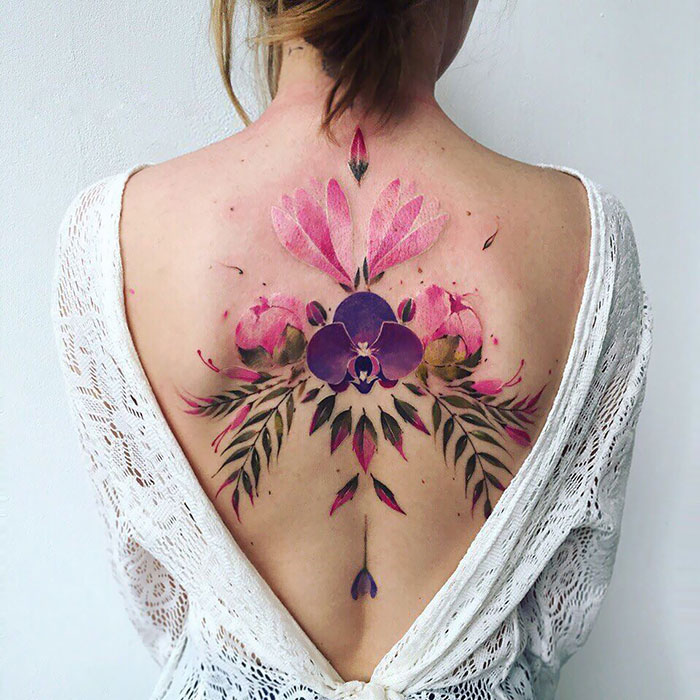 Floral back tattoo design with pink and purple flowers displayed on a woman wearing a white shirt.