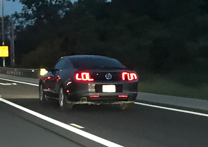 Funny car with LOL-shaped tail lights driving on a street at dusk.
