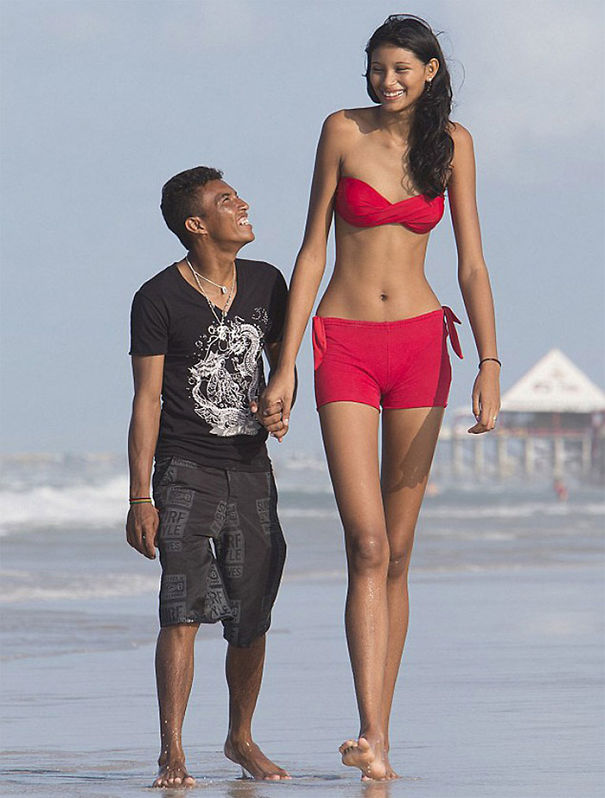 Tall woman in red outfit holding hands with shorter man walking on the beach, highlighting everyday struggles of tall people.