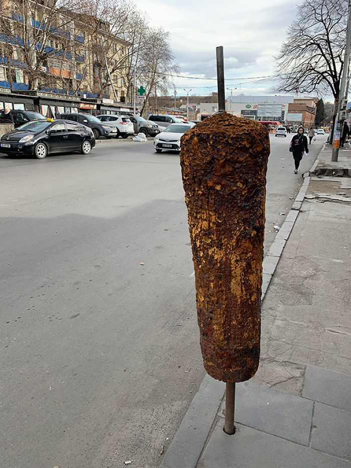 Rusty metal pole covered in layers of rust that looks like a delicious grilled meat skewer on a city sidewalk.