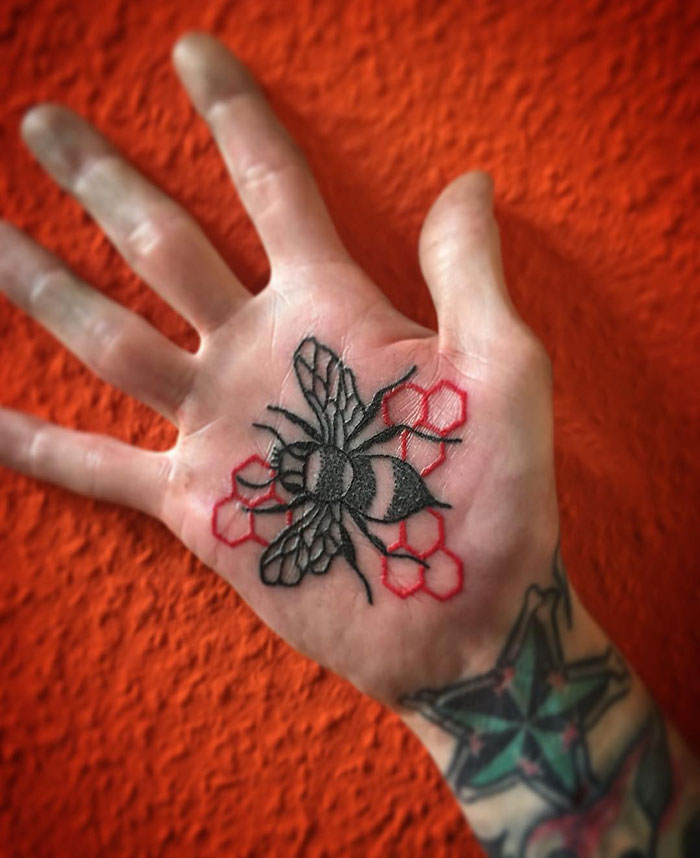 Bee design palm tattoo with red hexagon patterns on the hand against an orange background.