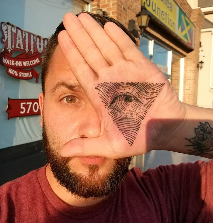 Man with intricate eye design palm tattoo, holding hand up to face outside a tattoo shop.