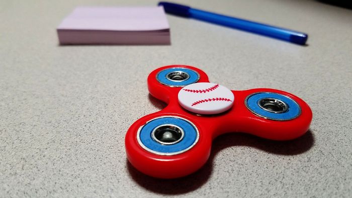 Red fidget spinner with baseball design, pen, and note pad on a gray desk highlighting forgotten hottest trends.