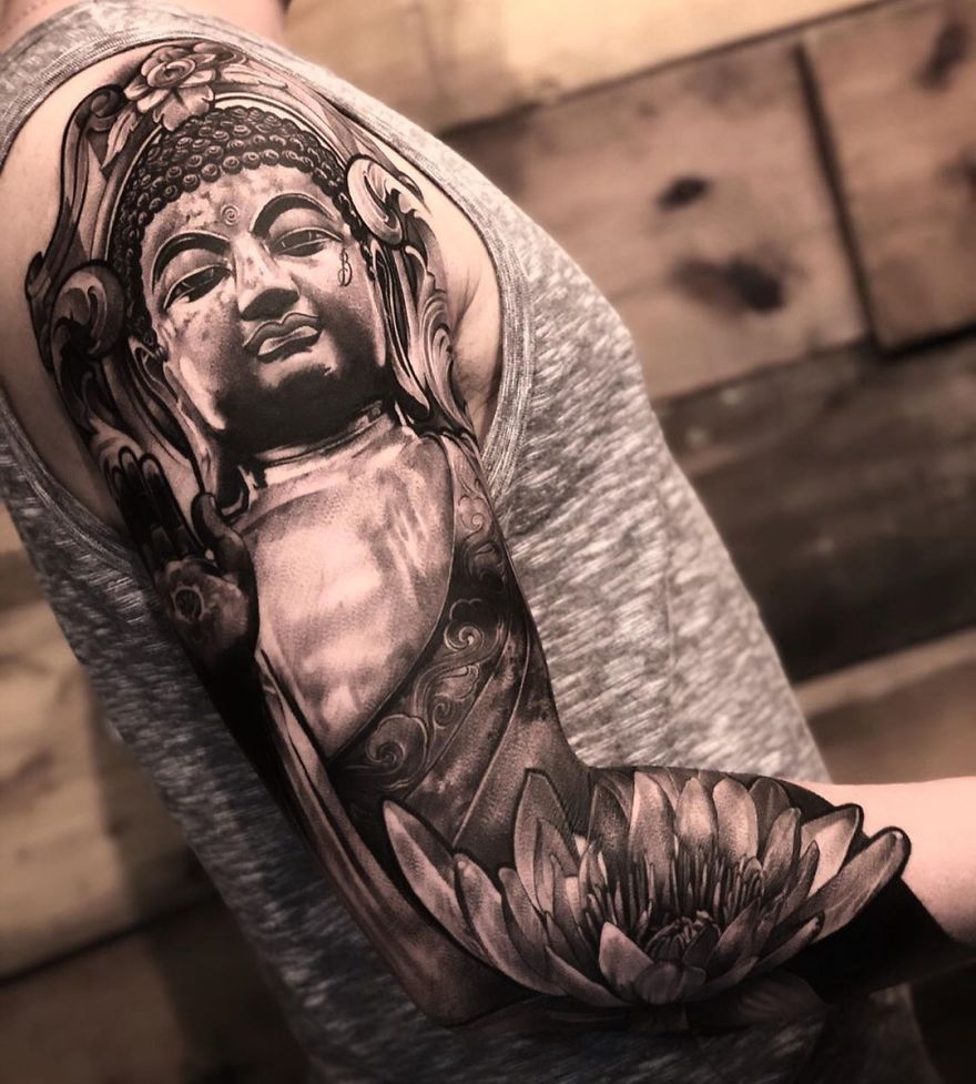 Buddha tattoo on upper arm in grayscale, showcasing masculine tattoo art with intricate details against a wooden background.