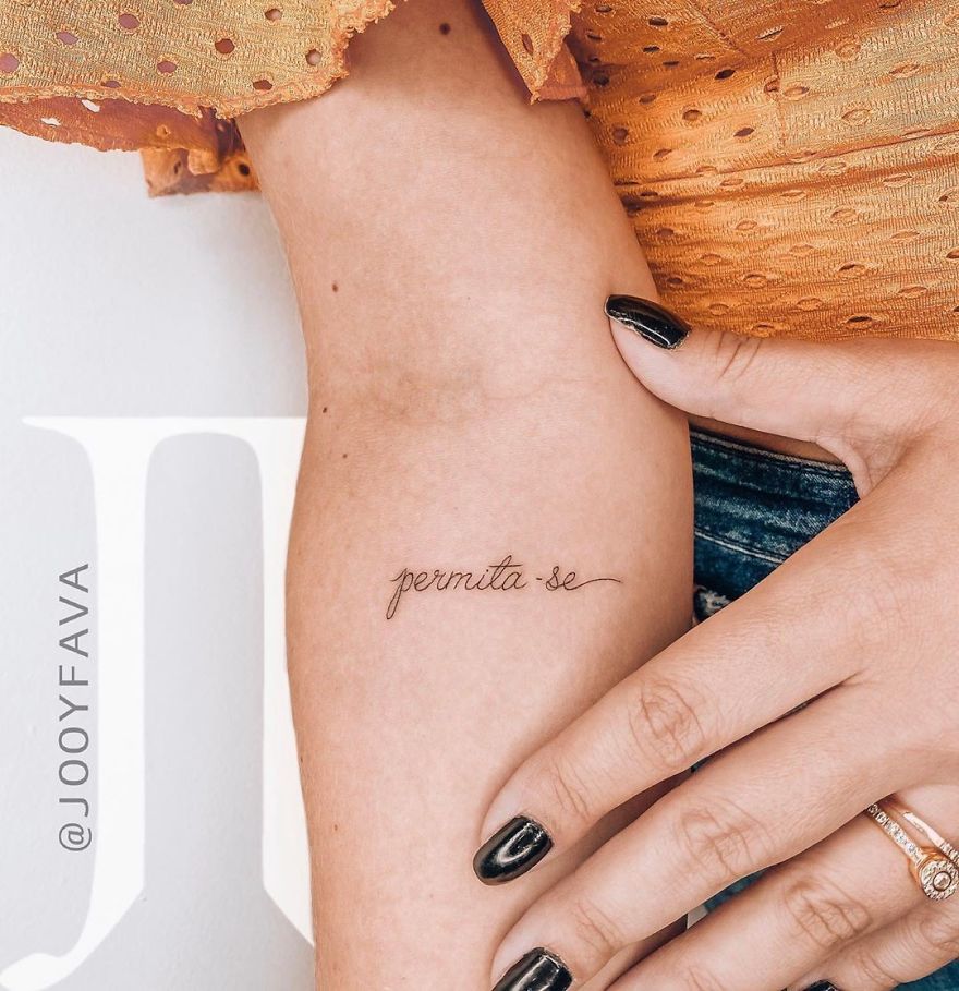 Close-up of a feminine tattoo of the words "permita-se" on an arm, with stylish black nail polish.