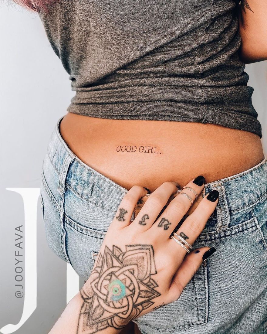 Hand with ring tattoos touching lower back with "Good Girl" text, demonstrating popular 2019 feminine tattoos.