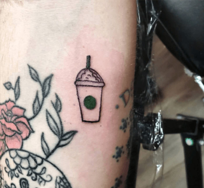 Tattoo of Starbucks cup on arm, surrounded by floral designs, symbolizing love for coffee.