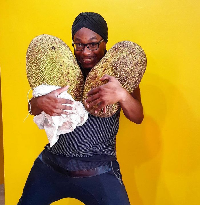 Man wearing glasses and a headwrap holding two large jackfruits with a joyful expression in a bright yellow room vegan hunters.